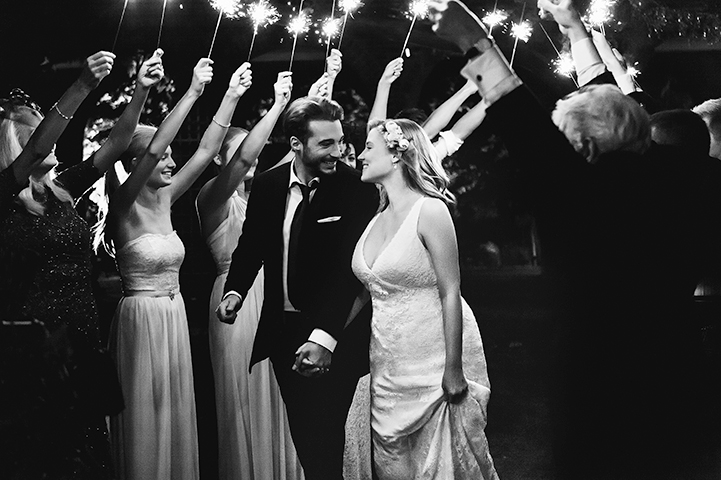 Couple in suit and wedding gown walking down aisle surrounded by people holding sparklers